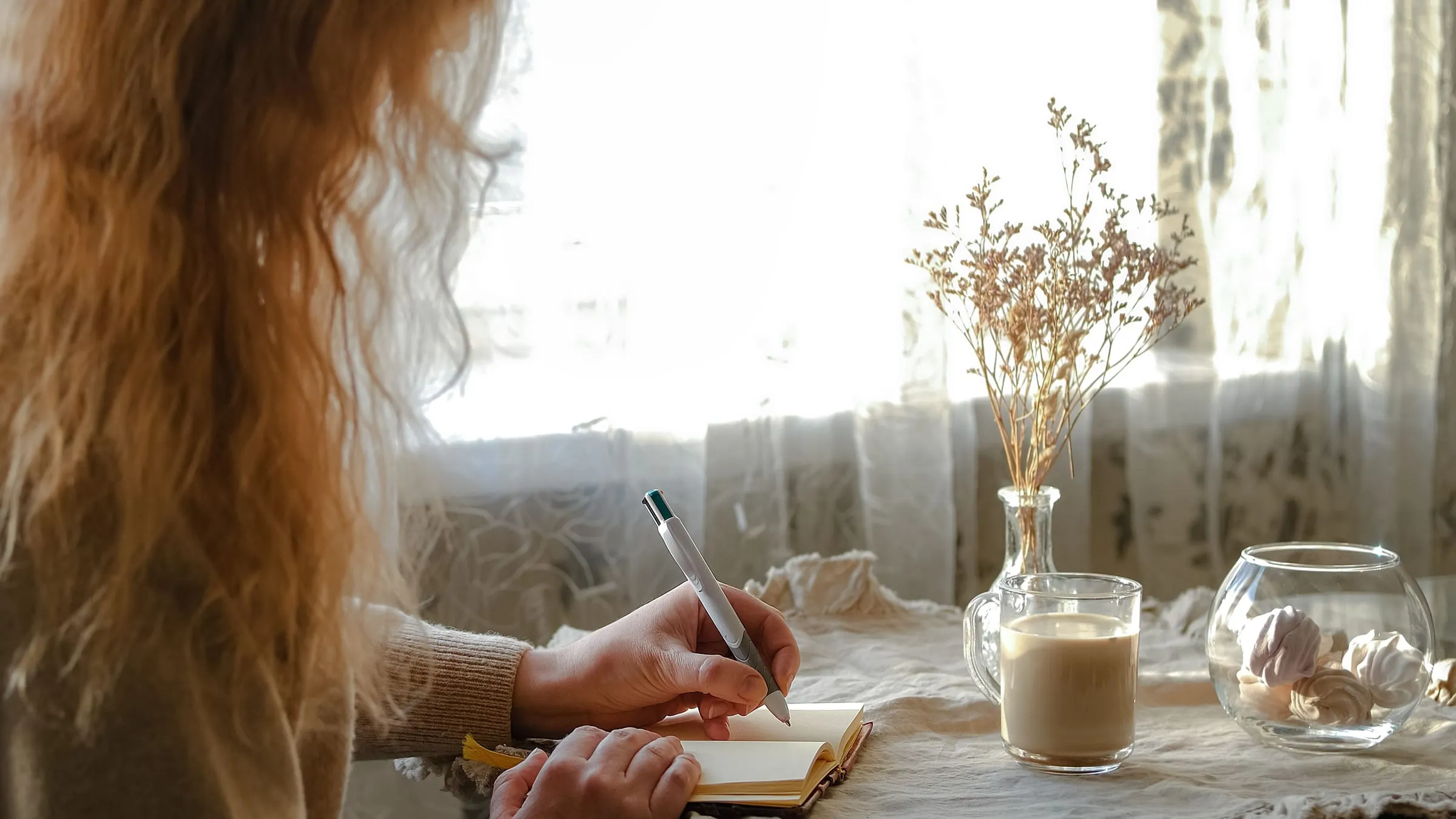 Who is my soulmate? A woman writes her answers in a notebook at a table by a lace-curtained window